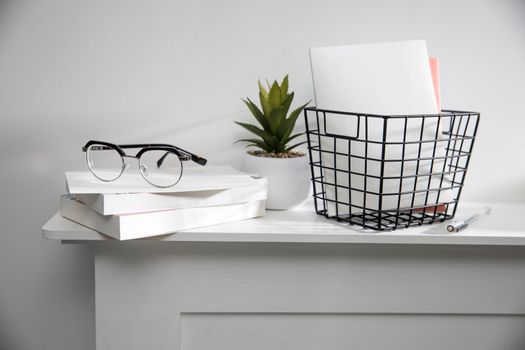 A stack of books, an artificial succulent plant in a ceramic pot, glasses, a metal basket with notebooks, a pen and a cup on a white table surface. Concept - office at home. Online training.