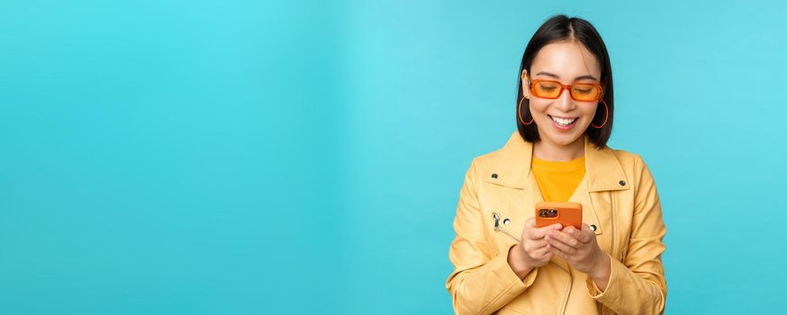 Smiling asian girl in sunglasses, using smartphone app, holding mobile phone, standing over blue background. Copy space