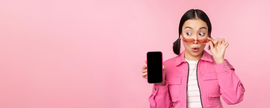 Stylish korean girl, young woman in sunglasses showing smartphone screen, mobile phone app interface or website, standing over pink background.