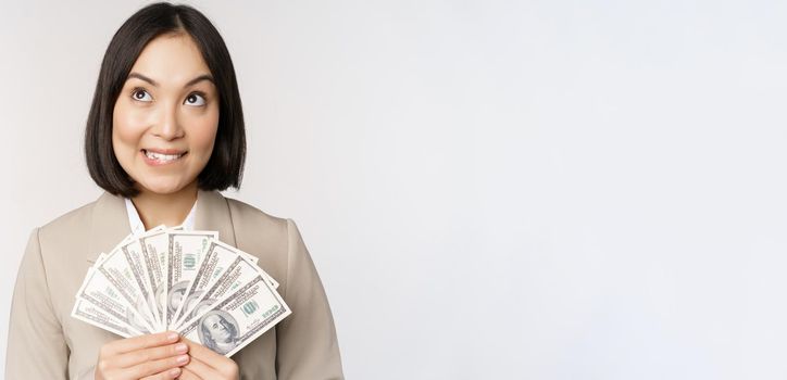 Image of asian corporate woman, happy businesswoman showing money, cash dollars and thinking, standing in suit over white background.