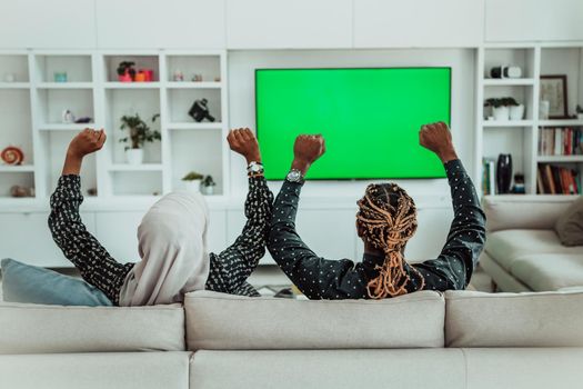 African Couple Sitting On Sofa Watching TV Together Chroma Green Screen Woman Wearing Islamic Hijab Clothes. High quality photo