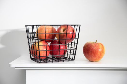 Five red apples in a black metal storage basket that stands on a white mantel. One apple is outside of basket. Copy space