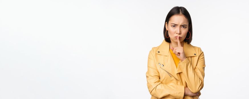 Portrait of angry korean girl shushing, woman frowning and tell to be quiet, showing hush, shh gesture, press finger to lips, taboo sign, standing over white background.