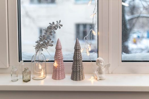 Gray and pink ceramic Christmas trees, a glass bottle with an artificial silver branch and a garland decorate the windowsill for Christmas.