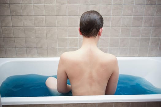 Unrecognizable girl with wet, smoothly combed hair sits in blue water in the bathroom with her back against the background of beige ceramic tiles