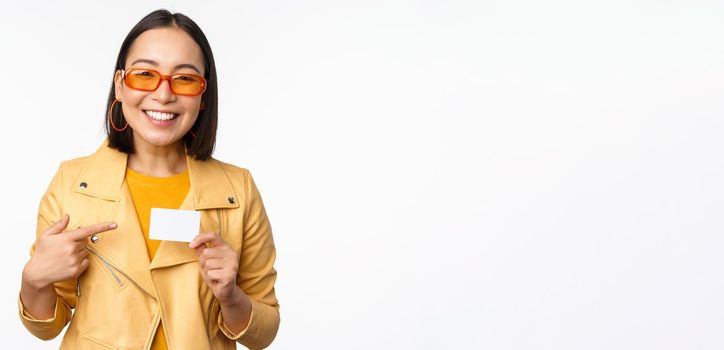 Portrait of beautiful modern asian girl in sunglasses, smiling happy, showing credit card, standing over white background. copy space