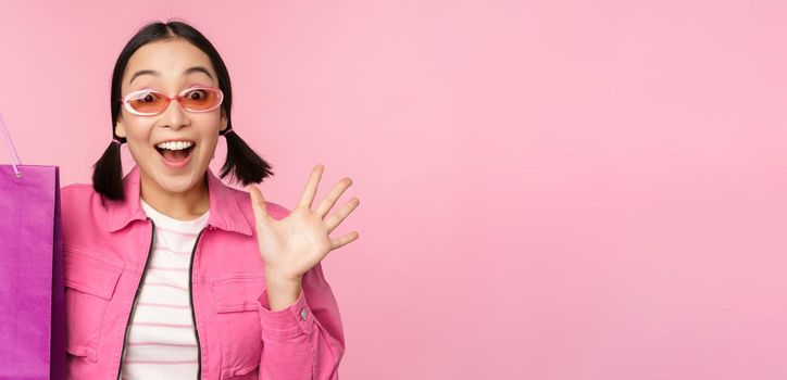 Shopping. Stylish asian girl in sunglasses, showing bag from shop and smiling, recommending sale promo in store, standing over pink background.