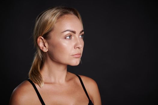 Beauty portrait of confident middle aged Caucasian blonde beautiful woman with clean healthy glowing tanned skin on black background.