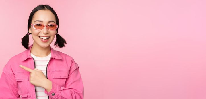 Portrait of smiling asian girl in stylish outfit, sunglasses, pointing finger left, showing advertisement, banner, standing over pink background. Copy space