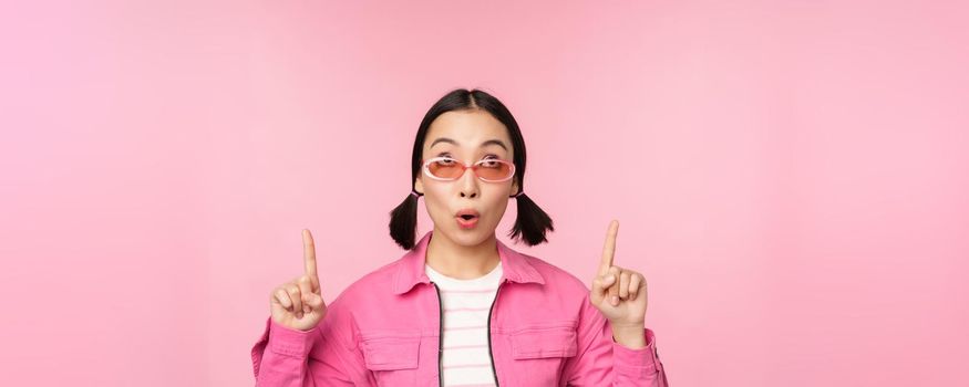 Close up portrait of asian woman gasping, looking surprised, pointing finger at banner, advertisement, standing over pink background.