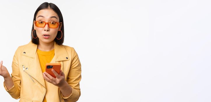 Portrait of korean girl in sunglasses, holding smartphone, looking confused and shrugging, standing over white background. Copy space