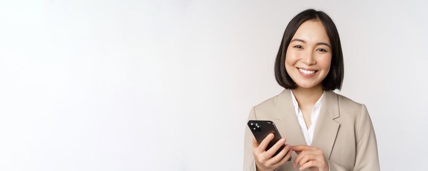 Close up portrait of korean woman, corporate lady in suit, using mobile phone and smiling, holding smartphone, standing over white background.