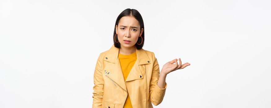 Image of frustrated korean woman, shrugging shoulders and raising hand, looking puzzled, cant understand smth, standing over white background.