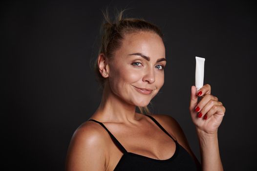 Beautiful Caucasian woman with tanned skin holding a tube of anti wrinkles smoothing anti puffiness under eyes cream, smiles looking at camera, isolated over black background with copy ad space