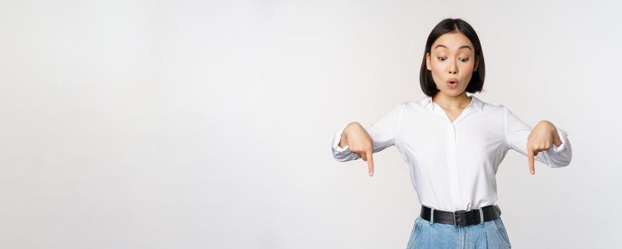 Surprised young asian female student, pointing fingers down and looking with amazed, impressed face expression, standing over white background.