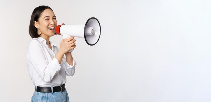 Image of young woman, korean activist, recruiter screaming in megaphone, searching, shouting at loudspeaker, standing over white background.