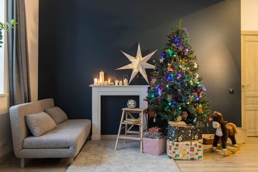 Home decoration before Christmas. Decorated Christmas tree with garland lights, rocking horse, gifts wrapped in elegant paper, artificial fireplace with a large white paper star and candles.