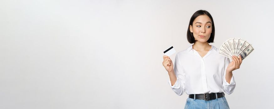 Image of asian woman looking at money dollars, holding credit card in another hand, thinking, standing over white background.