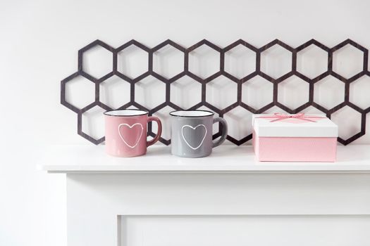 A pink and gray mug with a painted white heart, a white box with a red heart with a gift are located on the fireplace. Scandinavian style