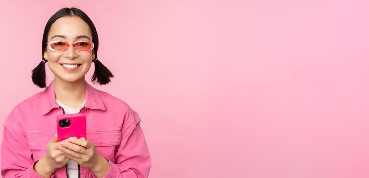 Portrait of korean girl in stylish sunglasses holding mobile phone, using smartphone app, standing over pink background. Copy space