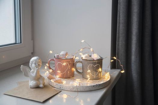 Apartment decoration for Christmas. Gray and pink mugs with painted white hearts with cocoa, marshmallows on tray on the window, composition decorates the interior on the eve of Christmas. Copy space