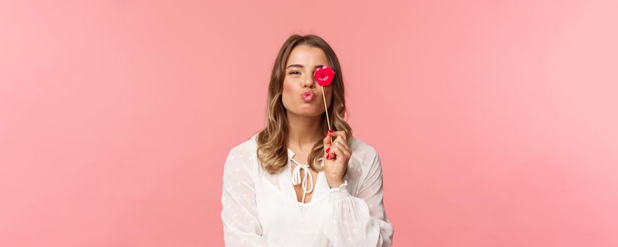 Spring, happiness and celebration concept. Close-up portrait of feminine lovely blond girl in white dress, folding lips in kiss and holding carton stick, partying having fun, stand pink background.