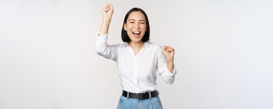 Image of happy lucky asian woman hooray gesture, winning and celebrating, triumphing, raising hadns up and laughing, standing over white background.