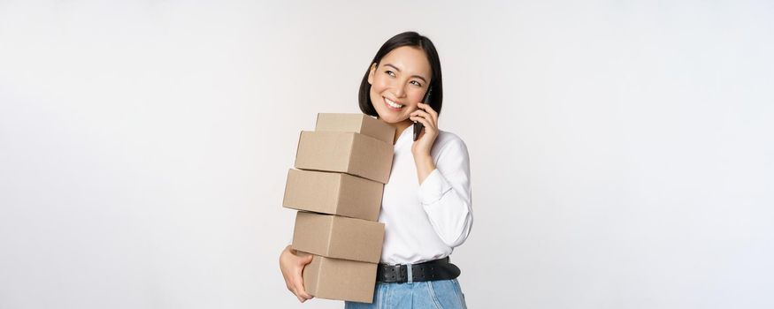 Image of young asian businesswoman answer phone call while carrying boxes for delivery, posing against white background.