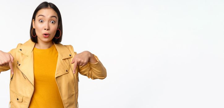 Portrait of surprised asian girl, looking amazed and impressed, pointing fingers down, showing banner with shocked face expression, white background. Copy space