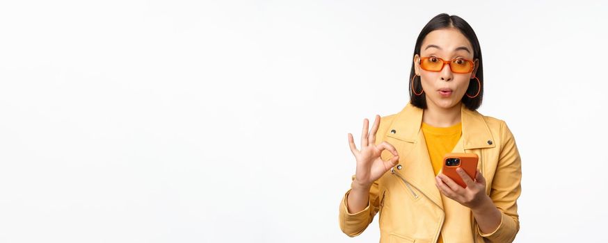 Amazed asian girl in sunglasses, showing okay, ok sign, holding smartphone, looking impressed, recommending smth, standing over white background.