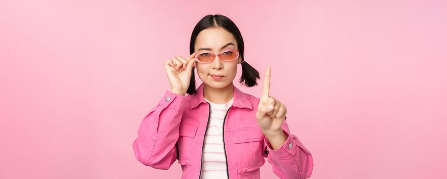 Image of asian girl with serious face, showing prohibit, scolding gesture, shaking finger pointing up, express disapproval, standing over pink background.