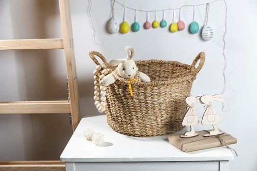 Interior decoration for Easter. A rag rabbit peeks out of a wicker willow basket on the table. A garland of wooden painted eggs above it.