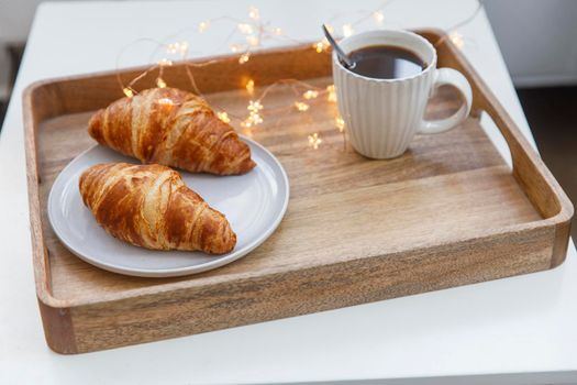 Freshly baked croissant on a gray round plate, white cup with coffee and garland on a tray on the table