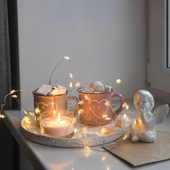 Apartment decoration for Christmas. Gray and pink mugs with painted white hearts with cocoa, marshmallows on tray on the window, composition decorates the interior on the eve of Christmas. Copy space