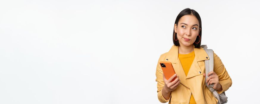 Asian girl traveller with backpack, holding mobile phone, using smartphone app, looking thoughtful, standing over white background.