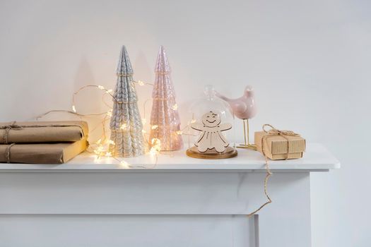 Christmas composition decorates the house. Ceramic figurines of gray and pink Christmas trees, a garland, gifts wrapped in brown paper, candles are located on a white chest of drawers. Copy space