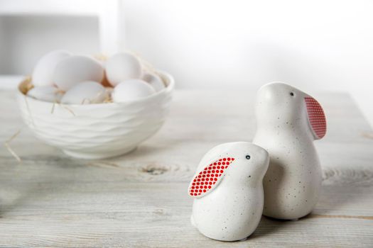 Two figurines of white hares with red ears big and small on a wooden saw cut and a bowl with eggs in the background in a white Scandinavian style kitchen