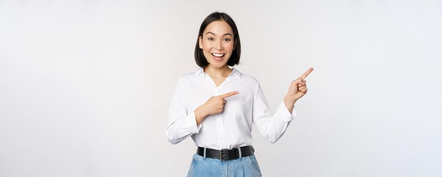 Portrait of beautiful asian woman, saleswoman pointing fingers right and showing info, banner or logo, sale advertisement, standing over white background.