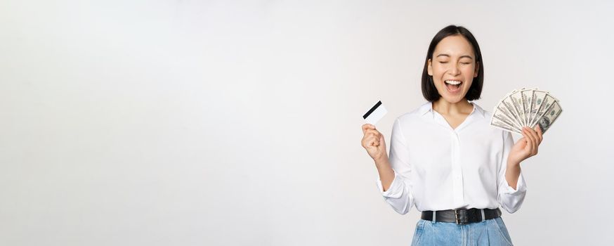 Finance and money concept. Happy young asian woman dancing with cash and credit card, smiling pleased, posing against white studio background.