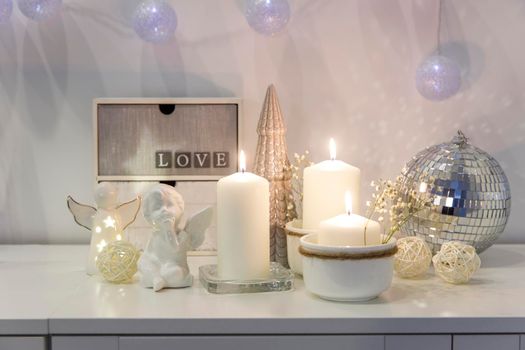 The concept of decorating the house for Christmas. Lighted candles, a garland, a disco ball, an angel figurine on a white chest of drawers. Place for text