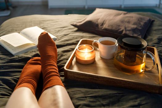 Soft photo of woman`s legs in the woolen socks on the bed with book and cup of tea and candle on the tray. Interior and home cosiness concept. Top view