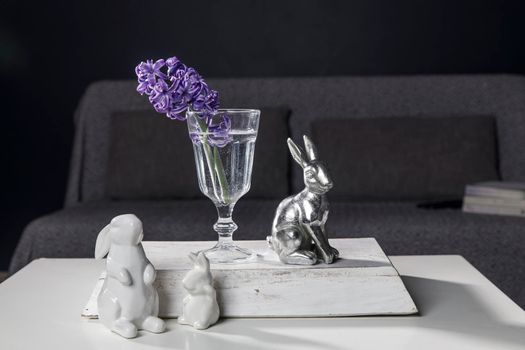 Ceramic figurines of Easter bunnies of different sizes on the table. Blue and white hyacinths in glass cups on a dark background. Easter design