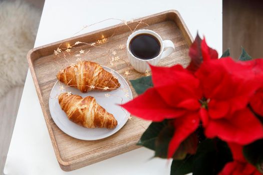 Freshly baked croissant on a gray round plate, a white cup of coffee and a garland on a tray on the table. Blooming poinsettia.