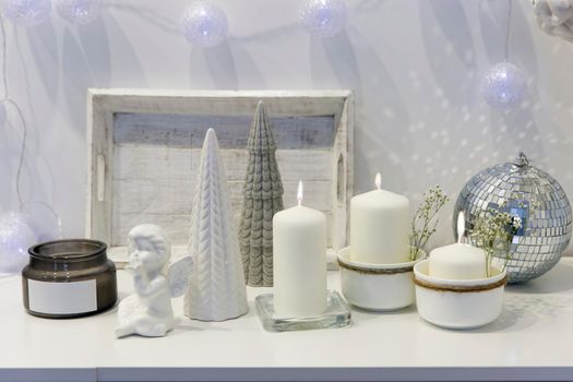 The concept of decorating the house for Christmas. Lighted candles, a garland, a disco ball, an angel figurine on a white chest of drawers. Place for text