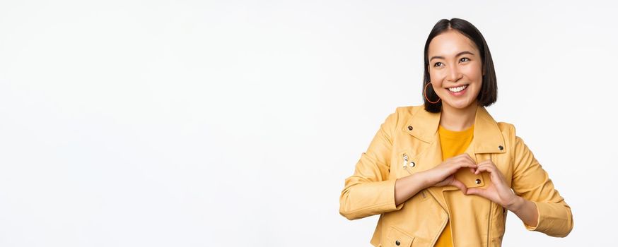 Image of beautiful asian girl showing heart, love sign, express care and romantic feelings, standing over white studio background.