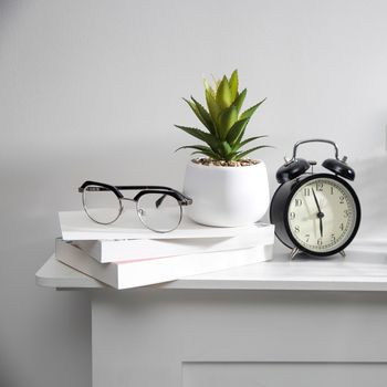 A stack of books, an artificial succulent plant in a ceramic pot, glasses, a metal basket with notebooks, a pen and a cup on a white table surface. Concept - office at home. Online training.