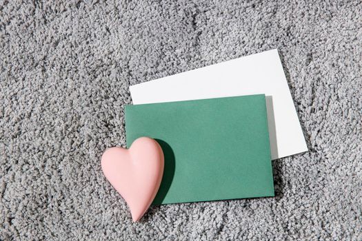 Greeting card for Valentine's Day. White and green envelopes, ceramic pink heart on a grey fluffy carpet background. Copy space. Place for text