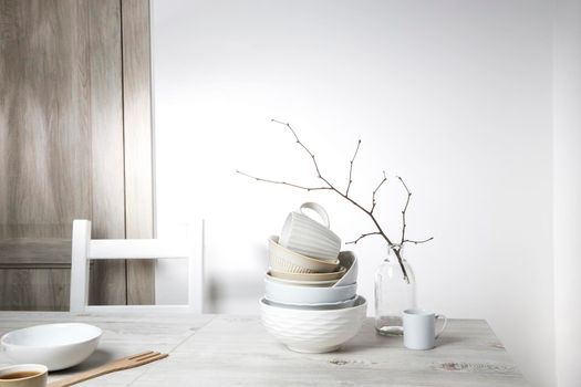 A pyramid of white bowls, plates and cups of different sizes and colors on a beige table in the kitchen. An unblown linden branch in a glass vase. Scandinavian style. Place for your text