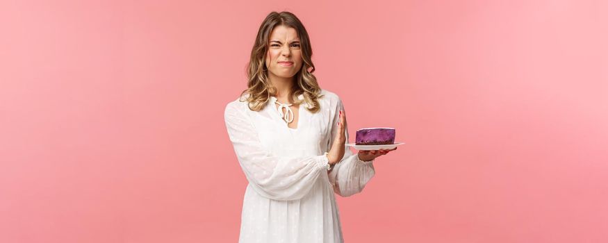 Holidays, spring and party concept. Portrait of dissatisfied and displeased blond girl in white dress rejecting, declining eat tasteless cake, grimacing and show stop sign at dessert, pink background.