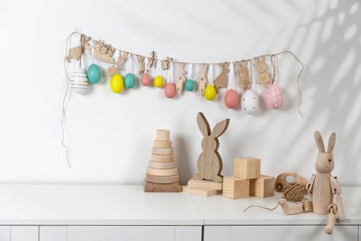 Fragment of the interior. Decorated children's room for Easter. A garland of plastic eggs and hares cut out of cardboard on the wall. Wooden rabbits and wooden cubes on the table. Place for your text. Easter card.